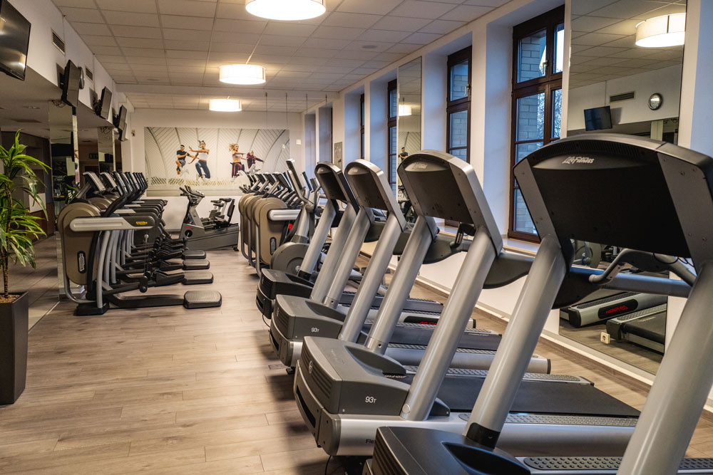 In einem Raum mit großen Fenstern stehen vier Laufbänder und dahinter Crosstrainer und Standfahrräder. An den Wänden hängen Spiegel und Fotos von Sportlergruppen.