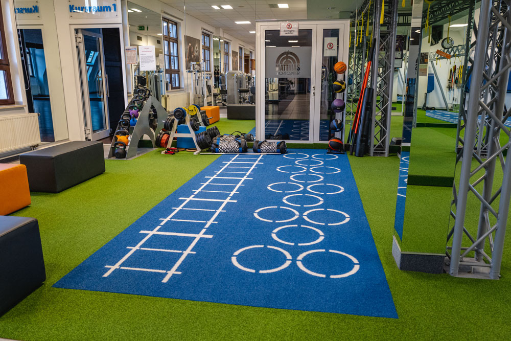 In einem Raum mit einem an Rasen erinnernden grünen Bodenbelag und einem blauen Bereich mit weißen Markierungen stehen an einer versiegelten Wand diverse, Fitnessgeräte wie Hanteln, Gewichtssäcke und Flexi-Bars.
