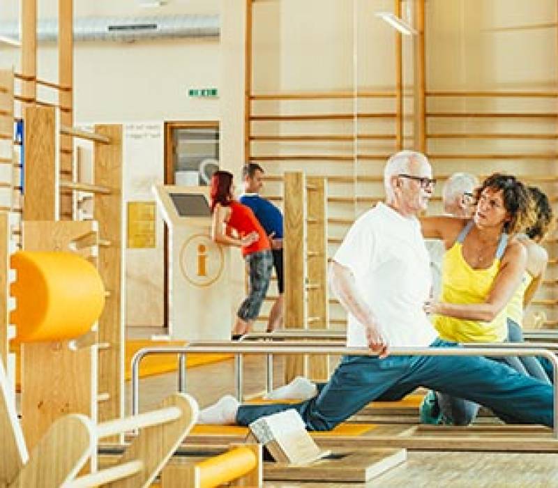 Ein &auml;lterer Mann wird im Five-R&uuml;cken- und Gelenkzentrum in einer &Uuml;bung korrigiert. Der Mann geht in die Spagatposition.