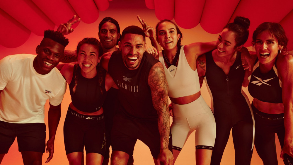 Eine Gruppe von sieben jungen Leuten unterschiedlicher Hautfarbe in Fitnesskleidung legen sich die Arme um die Schultern und lachen in die Kamera.