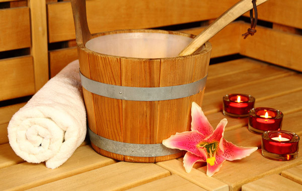 Ein hölzerner Aufgusseimer mit Kelle steht auf einer Sauna-Holzbank. Links daneben liegt ein zusammengerolltes weißes Handtuch. Eine exotische Blüte und drei Teelichter vervollständigen das Stillleben.